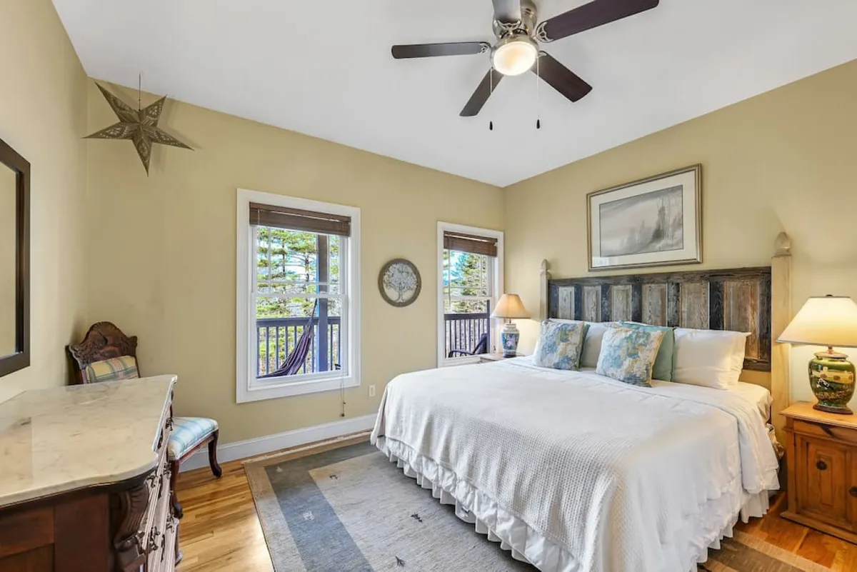 A king-sized bed with a rustic headboard is the focal point of this serene bedroom. Two windows on either side of the bed let in abundant natural light and offer scenic views. A decorative dresser adds functionality and charm to the space, while the calming tones and plush bedding evoke a restful retreat.