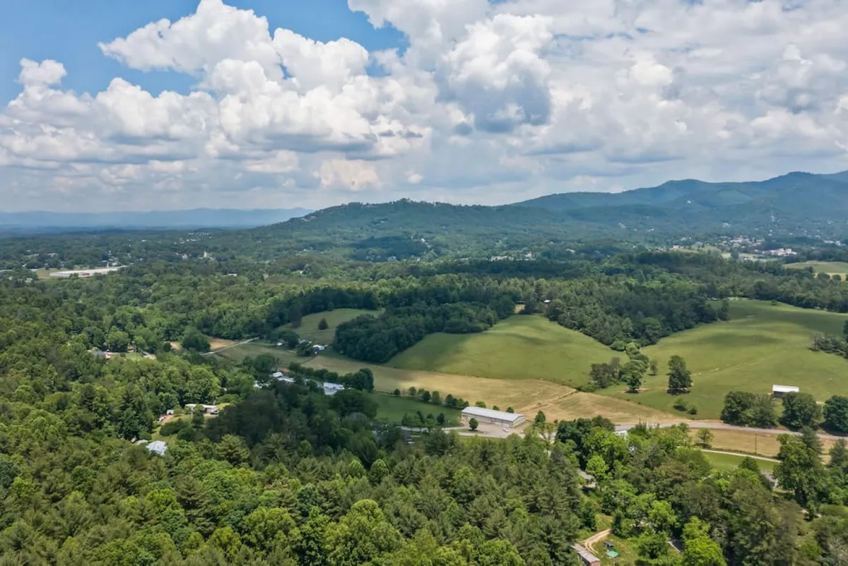 A wide view of rolling green hills, dense tree cover, distant mountains, and farmland beneath a cloudy sky. The scene radiates tranquility and captures the essence of the Blue Ridge region, with hints of hiking trails and open spaces for picnicking or nature exploration. The sunset glow drenches the landscape, inviting thoughts of outdoor adventures and scenic beauty.