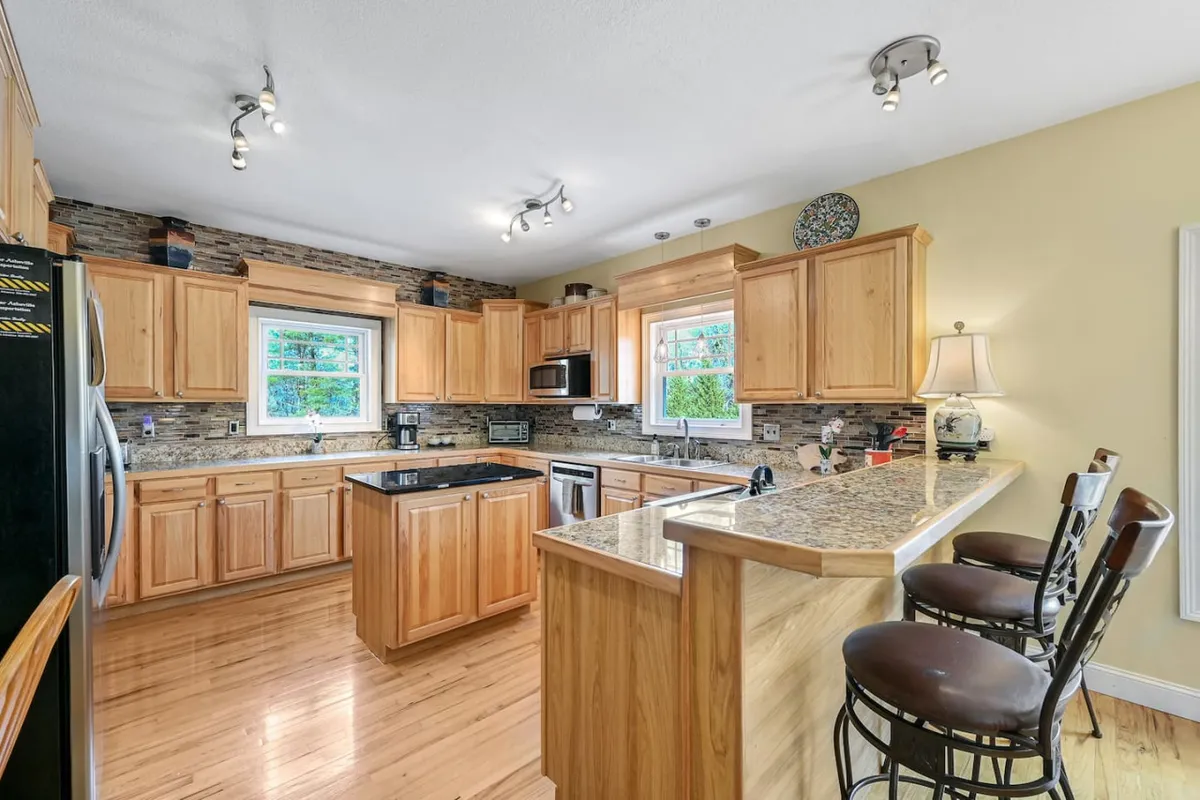 This image showcases a spacious kitchen with a central island, granite countertops, and warm wooden cabinetry. The scene is brightened by a window view, suggesting a welcoming and practical environment, ideal for cooking and entertaining.