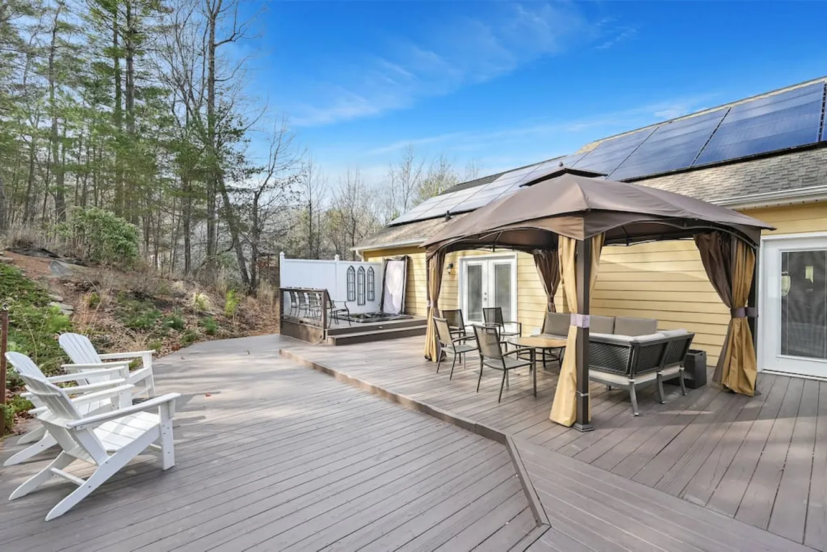 A covered patio area furnished with a dining table and chairs, set beneath a sturdy roof to provide shelter from the elements. The patio is adjacent to greenery, and solar panels are visible on the roof. The space is ideal for outdoor meals, morning coffee rituals, or socializing while immersed in nature’s tranquility.