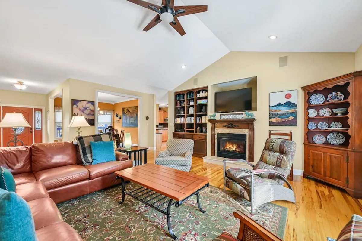 A cozy living room featuring a plush leather sofa, rustic wooden coffee table, inviting fireplace, built-in bookshelf adorned with charming decor, and tasteful artwork on the walls. The warm, welcoming ambiance is perfect for relaxing, gathering with family, or enjoying quiet evenings by the fire.