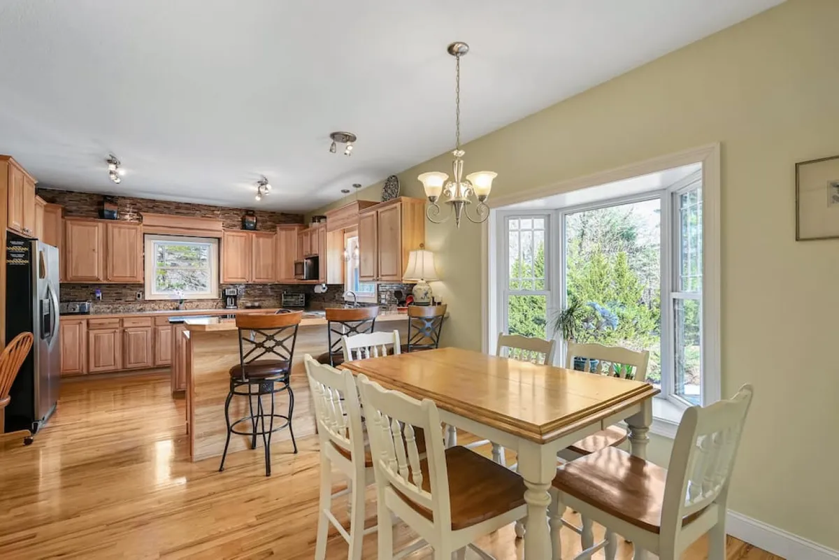 A cozy, sunlit dining area with a large wooden table, kitchen island, and wooden cabinets. A large window brings in plenty of natural light, and the space is perfect for shared meals or friendly gatherings.