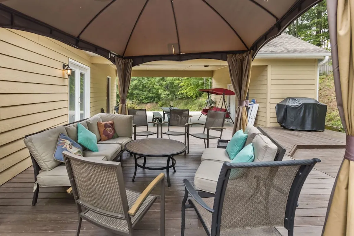 An outdoor seating area under a charming gazebo, featuring a round table in the center and surrounded by lush greenery. An outdoor grill is adjacent, hinting at space for barbecues and gatherings. The scene feels cozy and social—perfect for family get-togethers after a day out.
