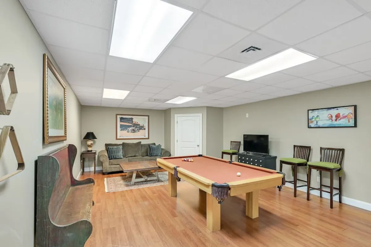 A playful, inviting game room featuring a pool table, comfortable sofa, coffee table, TV, and bar stools. The space is designed for both spirited competition and relaxed movie nights.