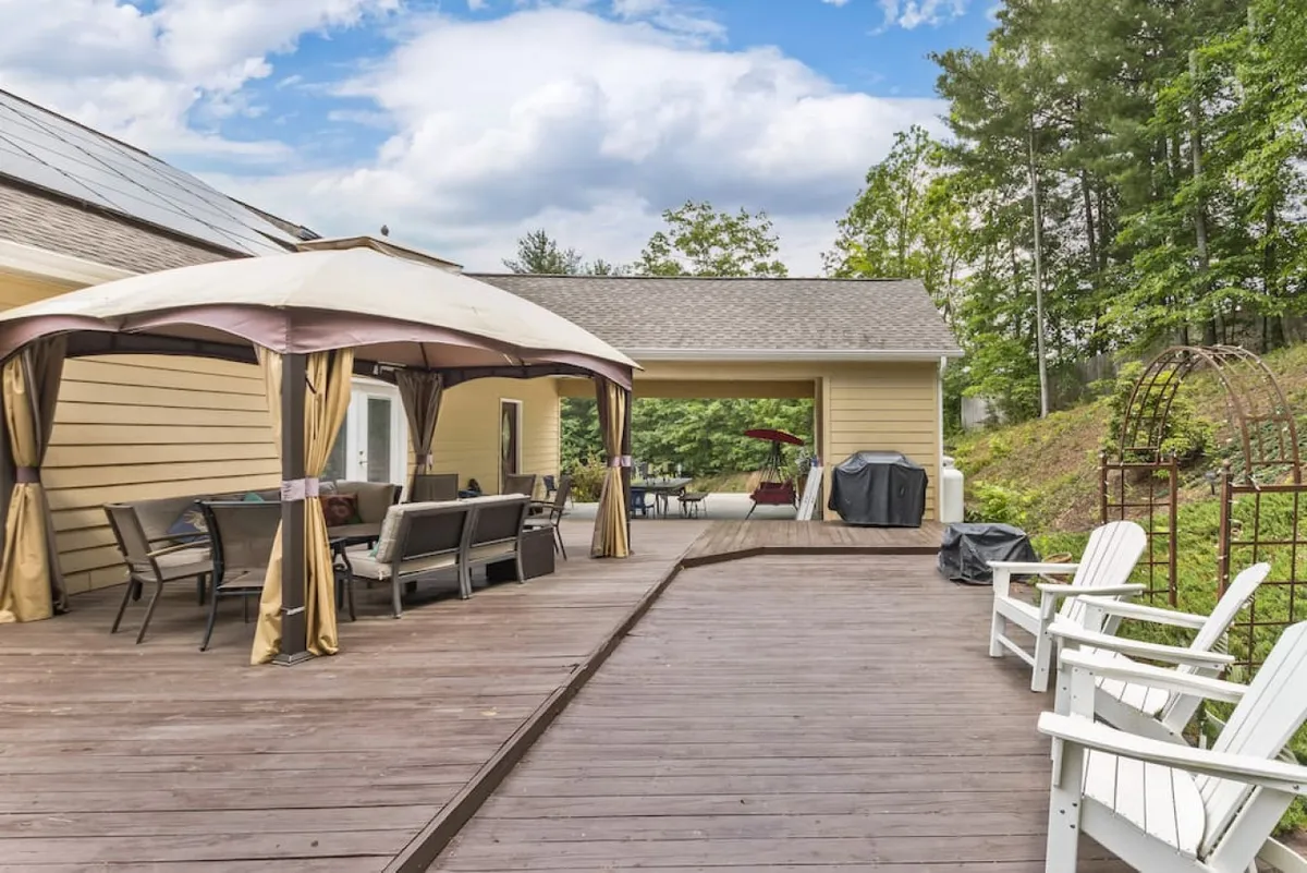 A covered deck area with a large outdoor dining table, grill, wooden deck flooring, and Adirondack chairs. The space is designed for outdoor dining and entertaining, ideal for sunset dinners or starlit evenings with friends.