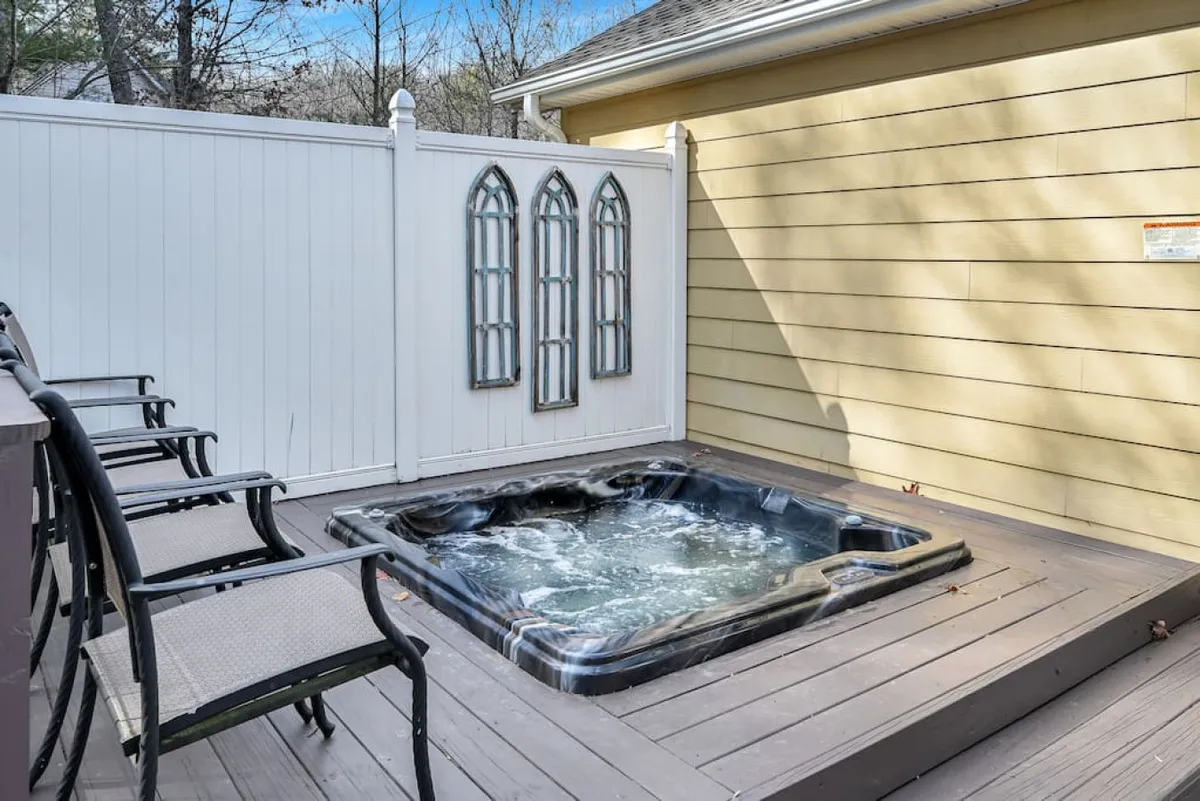 A tranquil corner of the deck featuring a built-in hot tub, bordered by decorative windows and surrounded by comfortable outdoor seating. The area is designed for relaxation, with easy access to both sun and shade, and a view out over the surrounding trees. This setting is ideal for unwinding after outdoor activities or enjoying a glass of wine in the evening.