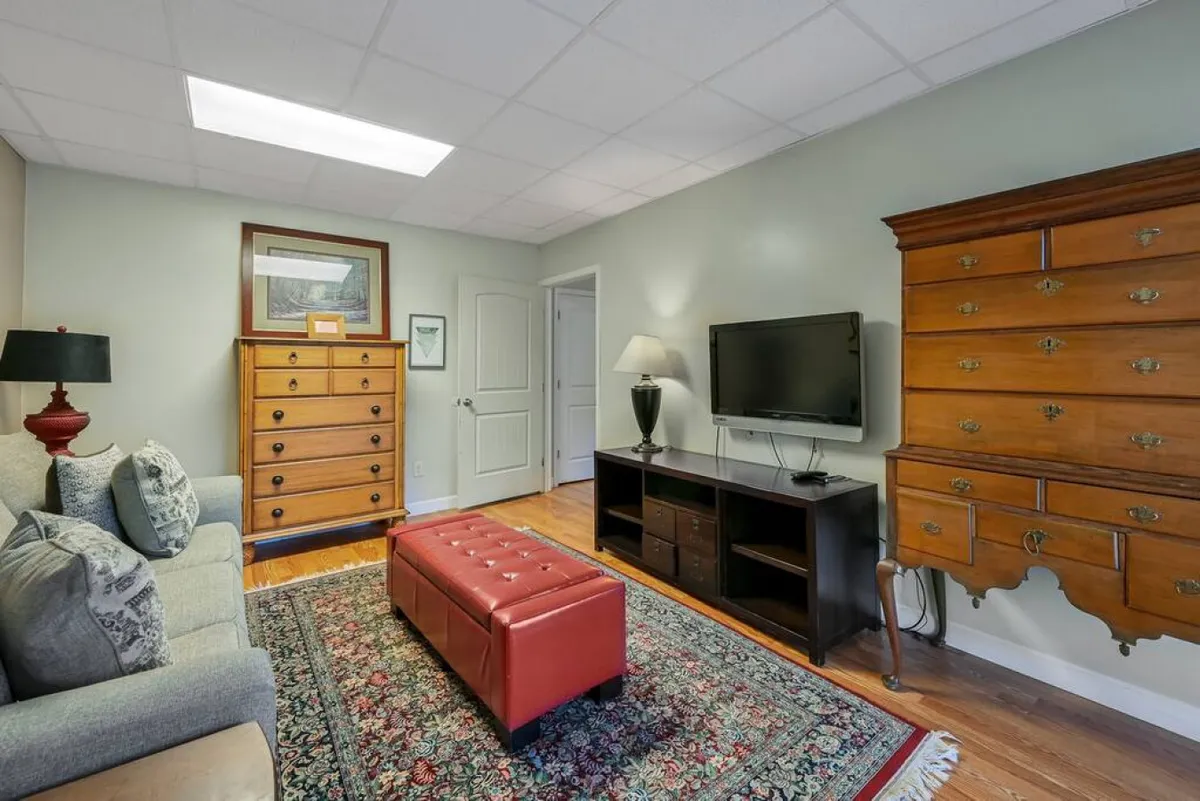 A comfortable bonus room featuring a sofa with patterned cushions, a red leather ottoman, and wooden dressers. The room is arranged for relaxation, with warm, homey touches that make it ideal as a hideaway for kids, teens, or additional guests. The decor and layout support flexible use for rest or play.