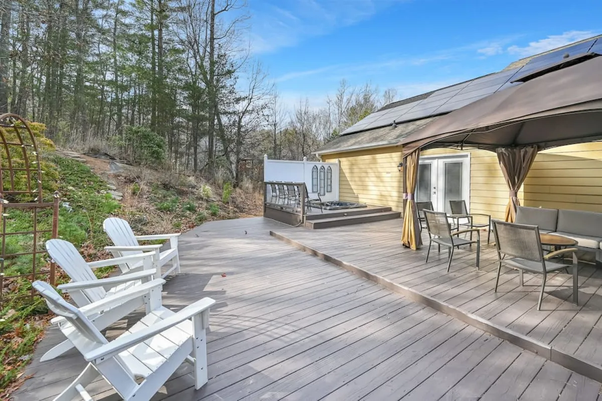 A spacious wooden deck set against a lush forest backdrop. White Adirondack chairs are spread across the deck, while a patio table and chairs sit beneath a gazebo. The surrounding trees provide both privacy and a calming natural ambiance, making the space ideal for group gatherings or peaceful solitude. Evening string lights suggest a space perfect for stargazing or sunset dinners.