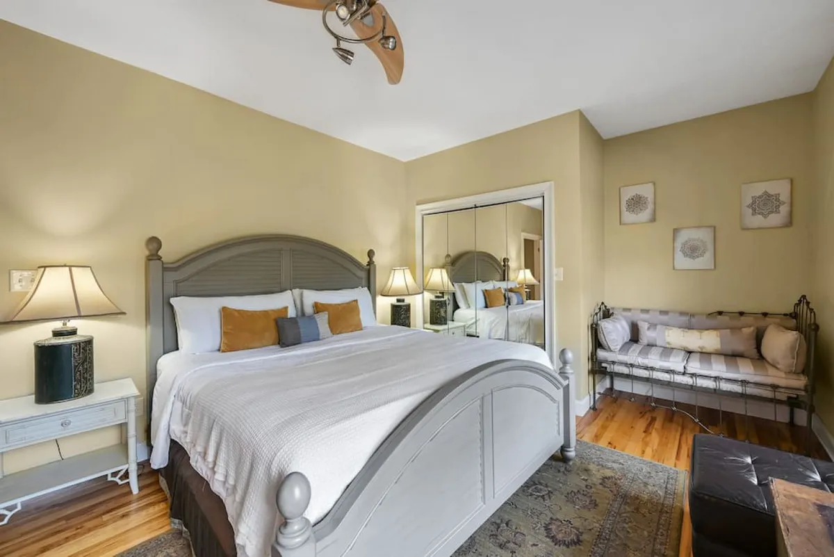 This tranquil bedroom features a bed with decorative pillows, a mirrored wardrobe, bedside lamps, and a seating bench. Soft natural light filters through the windows, illuminating the cozy nook and enhancing the room's comfort and style. The blend of ample storage and inviting decor underscores the retreat-like feel.