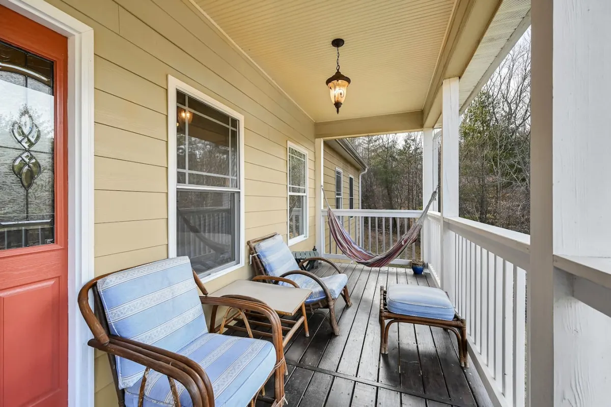 A cozy porch features a swing, rattan chairs, a hammock, and a view of the front door framed by lush greenery. The welcoming space is perfect for enjoying the outdoors, reading, sipping coffee, or socializing in a relaxed, casual setting.