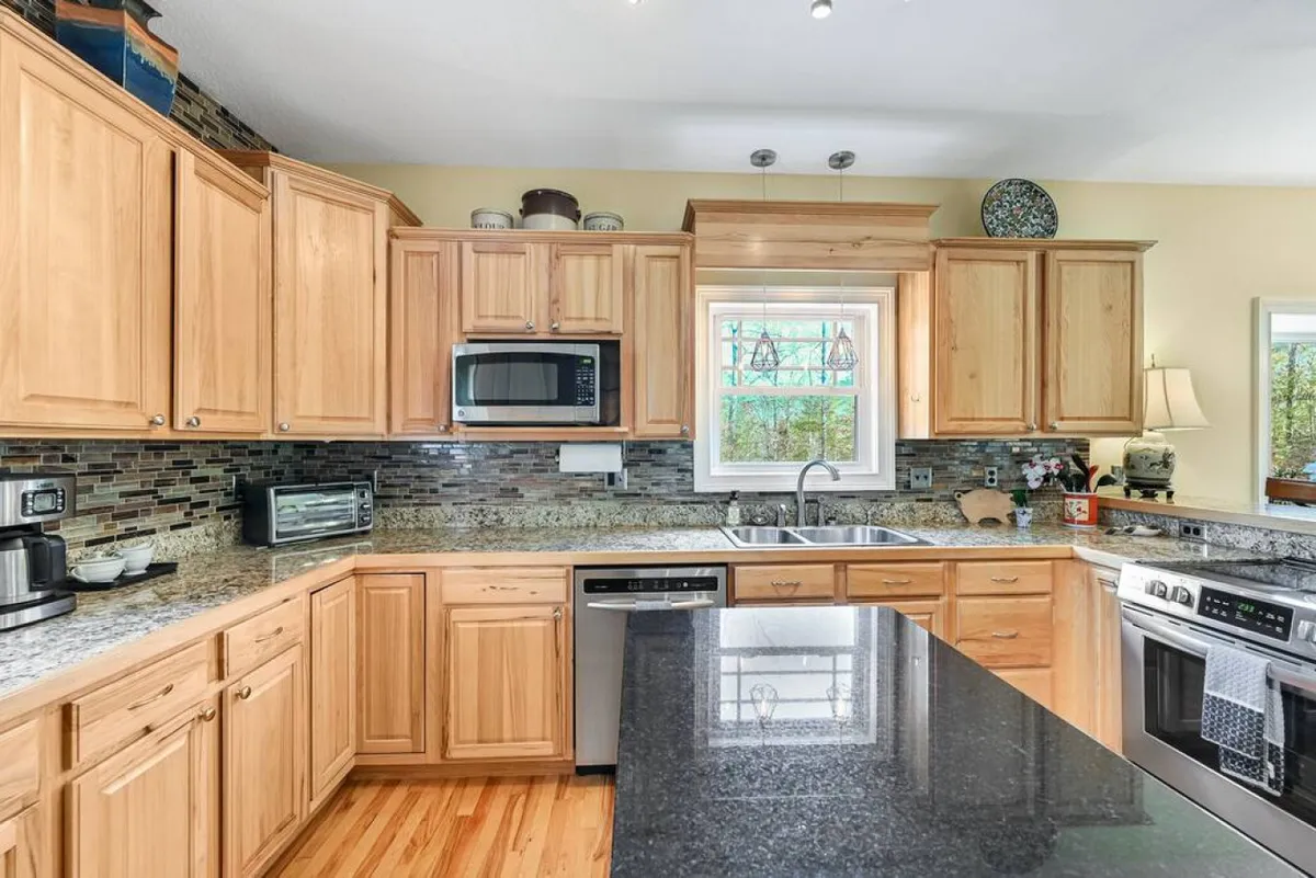 A cozy kitchen area with granite counters, wood cabinetry, stainless steel appliances, and a window bringing in natural light. Decorative touches on the shelves give a lived-in, welcoming feel ideal for both everyday cooking and special gatherings.
